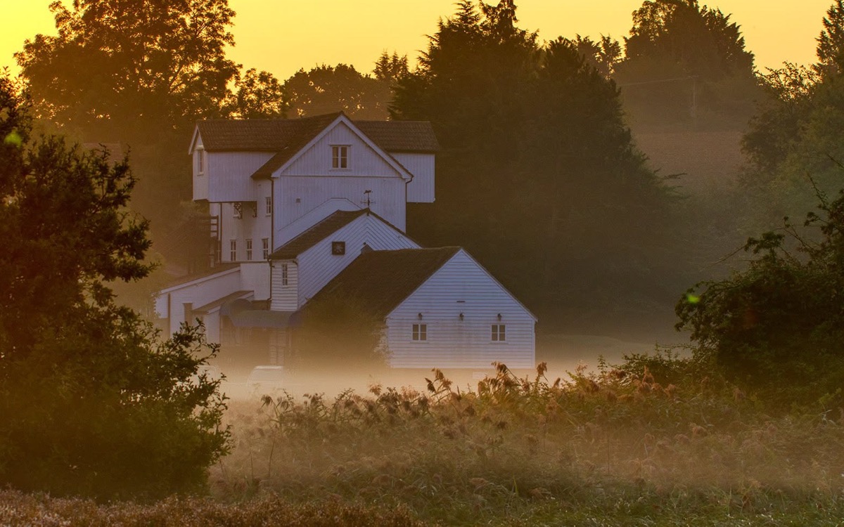 A weatherboarded mill above the water meadows at dawn, with mist lying across the fields